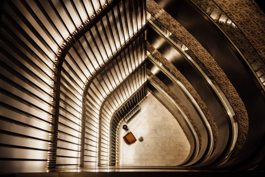Overhead view of a multi-story staircase with curved railings and illuminated steps, leading down to a chic seating area—a hidden gem often found in luxurious travel destinations—featuring a chair and two round tables on a marble floor.