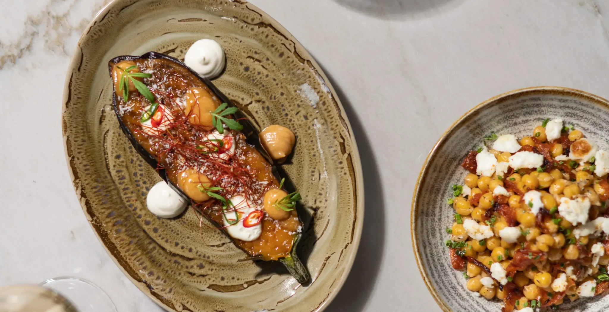 Ein ovaler Teller mit einer gebratenen Aubergine mit Soße, Sahnetupfen und Kräutern, daneben eine Schüssel Kichererbsensalat mit zerbröckeltem Käse.