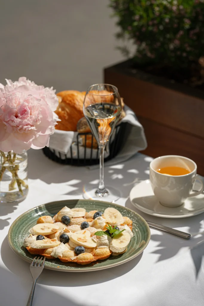 A plate of pancakes topped with banana slices, blueberries, and cream sits on a table at Donnersmarkt Restaurant, alongside a cup of tea, sparkling wine, bread in a basket, and a vase of pink flowers.
