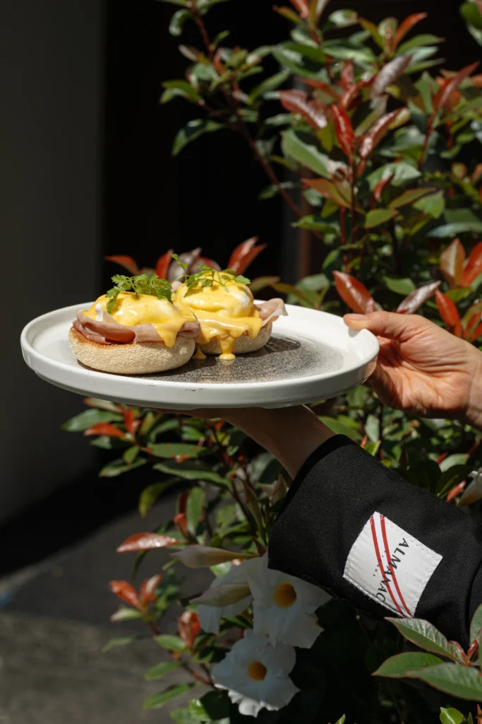 A person holds a plate with eggs Benedict topped with hollandaise sauce and herbs at Donnersmarkt Restaurant, set against a background of leafy plants.