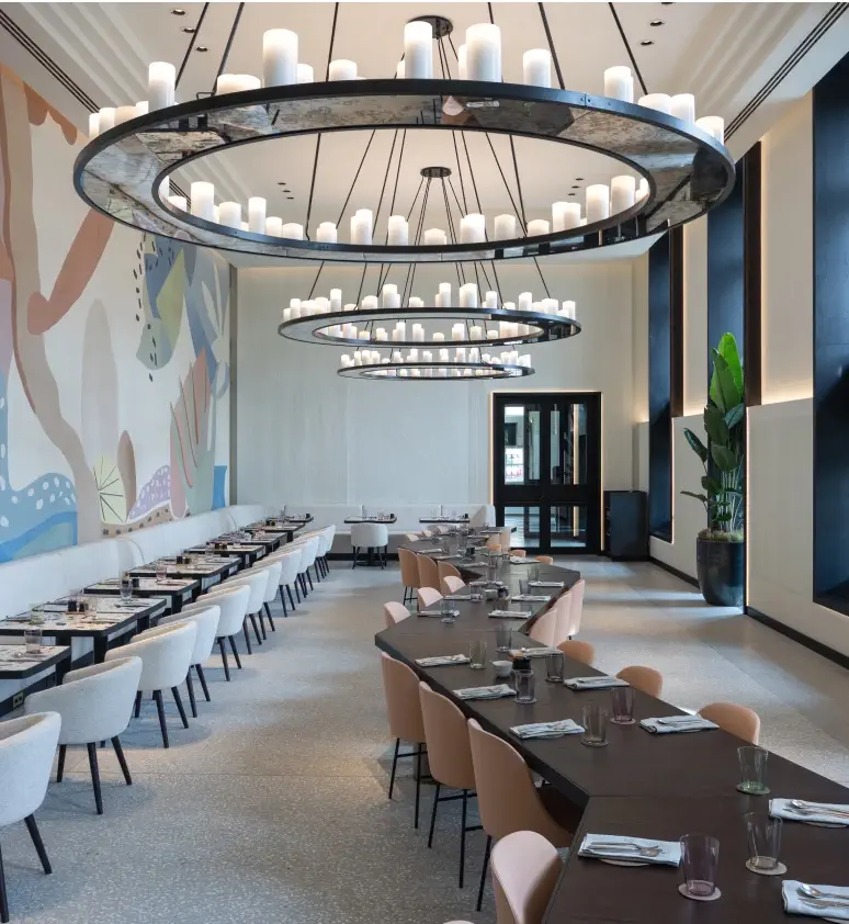 Modern dining area at Donnersmarkt Restaurant with long tables, cushioned chairs, large circular chandeliers, tall windows, a potted plant, and a colorful abstract mural on one wall.
