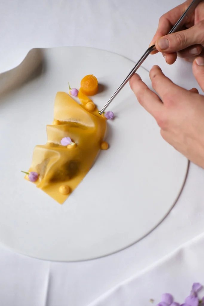 A hand uses tweezers to place a garnish on a plated dish of folded pasta with yellow sauce and purple edible flowers at Donnersmarkt Restaurant, beautifully arranged on a white plate.
