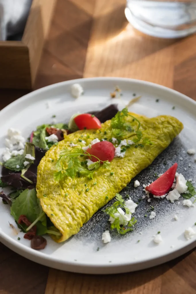 A yellow omelette folded over mixed greens, garnished with tomato wedges, crumbled white cheese, and herbs on a white plate at Donnersmarkt Restaurant.