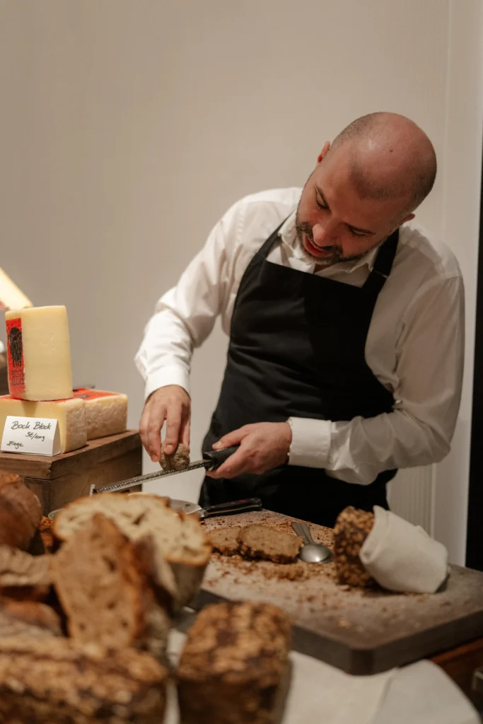 At Donnersmarkt restaurant, a man in a black apron slices bread on a wooden board, surrounded by assorted loaves and a large block of cheese.