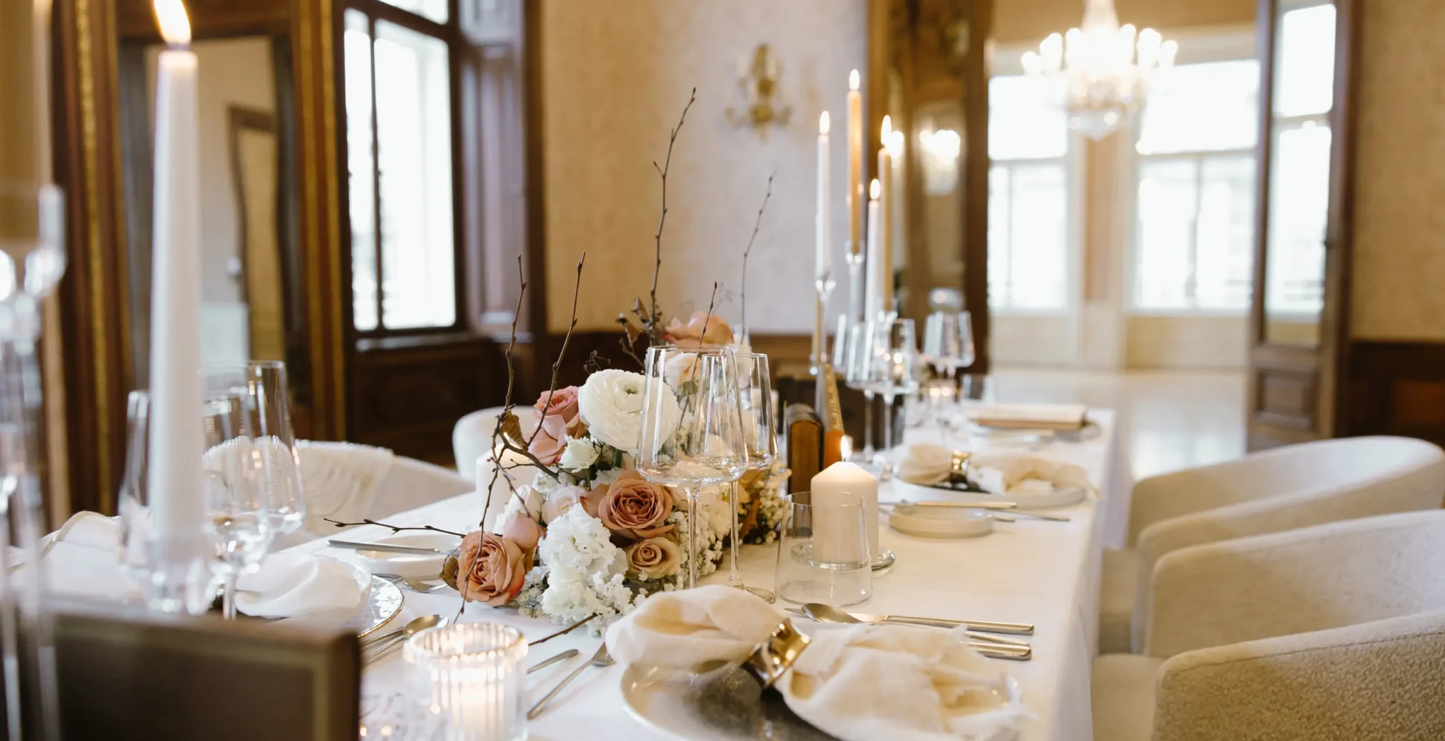 Elegant gedeckter Esstisch mit weißer Bettwäsche, floralen Tafelaufsätzen, Kerzen, Gläsern und Silberbesteck in einem hellen, gehobenen Raum mit großen Fenstern und Kronleuchtern.