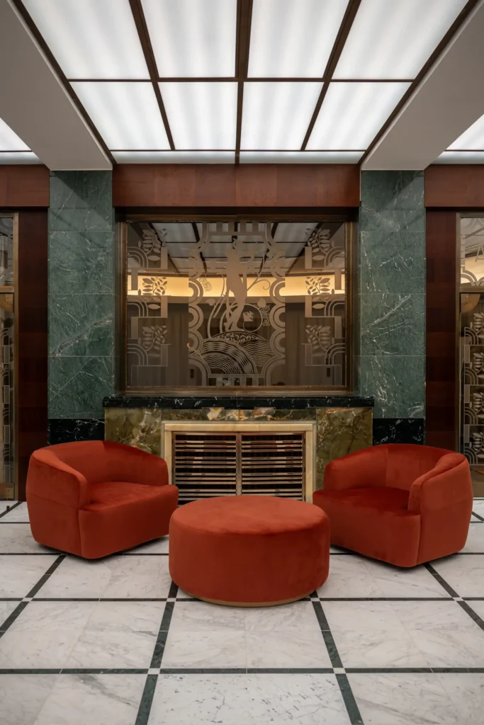 Three rust-colored velvet chairs and an ottoman form a cozy semi-circle on a white marble floor, creating a chic lounge area in front of a green marble fireplace—an inviting destination beside the decorative glass wall.