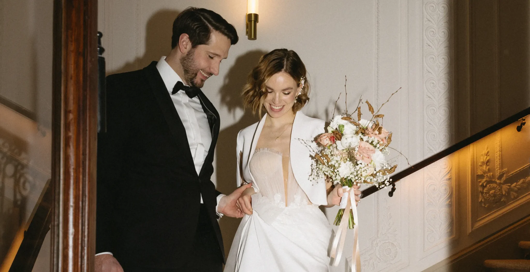 A bride and groom dressed formally descend a staircase together; the bride holds a bouquet of flowers and both appear happy.