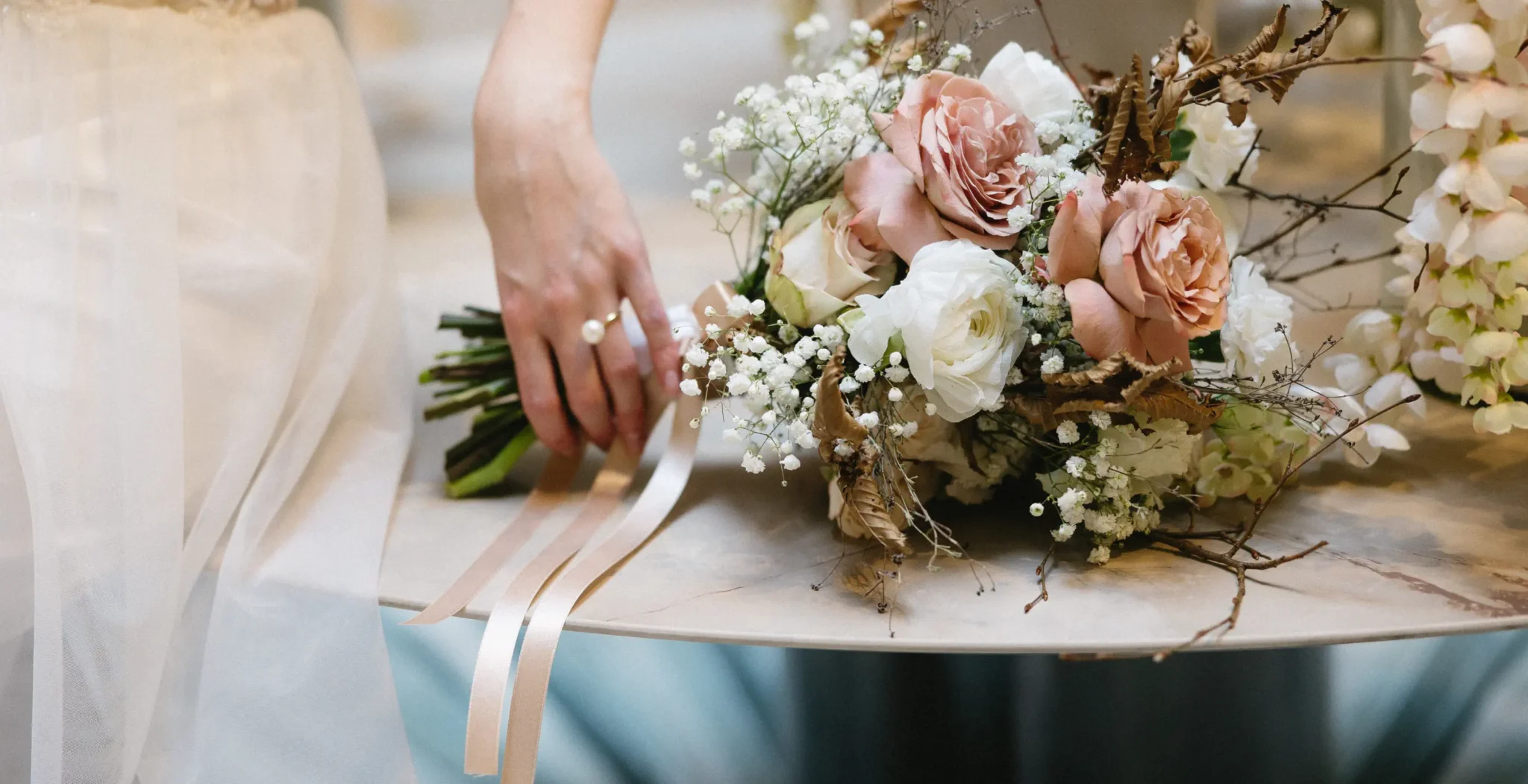 A person in a white dress reaches for a bouquet of pink, white, and beige flowers resting on a table.
