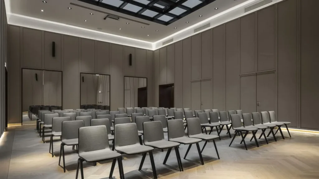 A modern conference room designed for events and meetings, featuring rows of empty gray chairs facing the front, high ceilings, and neutral-colored walls with recessed lighting.