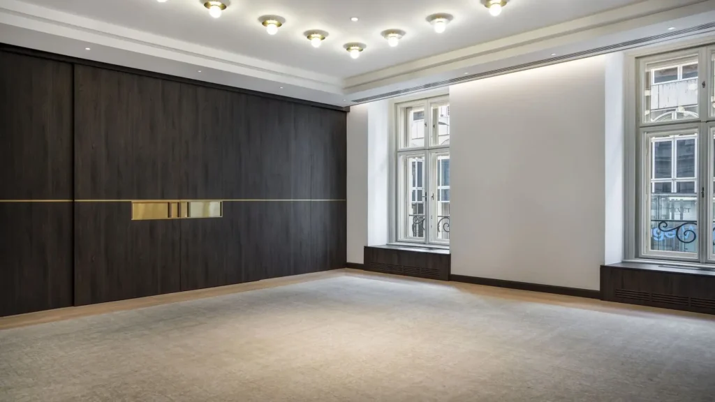 A modern, empty room with large windows, a beige carpet, dark wood paneling on one wall, and ceiling lights.