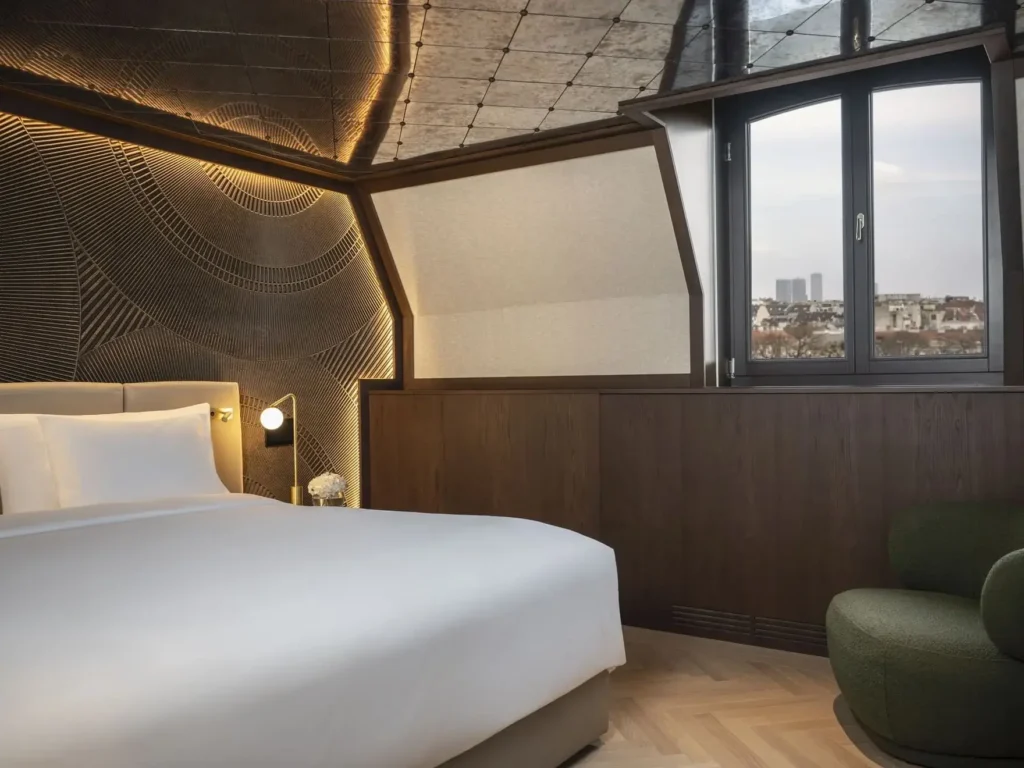 Experience the Almanac Park Suite: a modern hotel room featuring a large bed, textured accent wall, wood paneling, a city-view window, and a stylish green armchair.