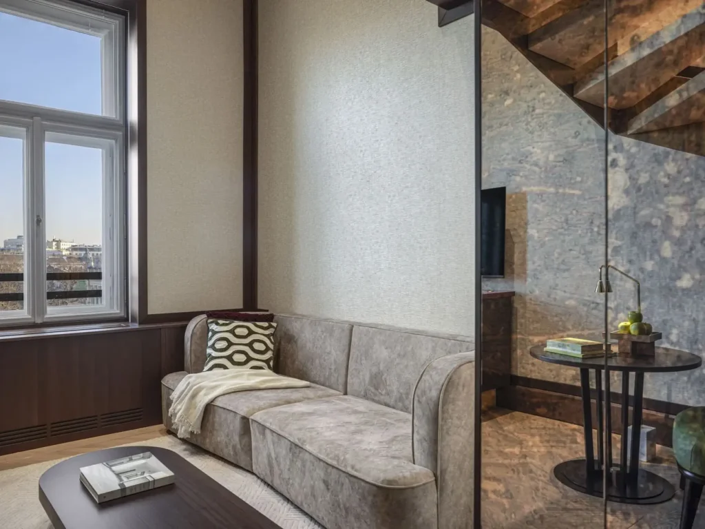 A modern Park Suite living room with a beige sofa, patterned pillow, throw blanket, coffee table with books, large window, and a glass partition revealing a small round side table in this stylish duplex.