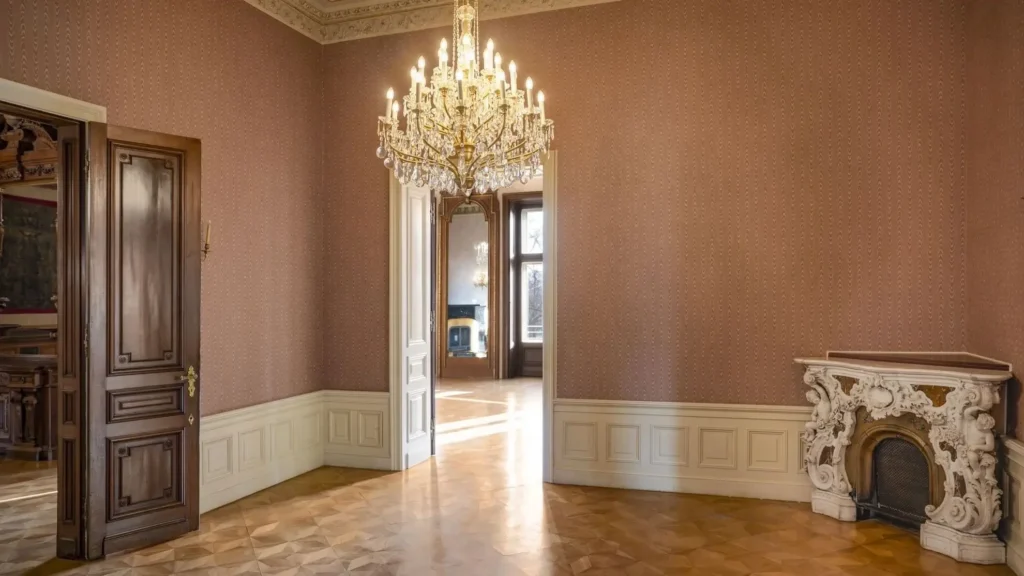 A chandelier hangs from the ceiling in an empty, ornate room with wood floors, patterned wallpaper, a decorative fireplace, and double doors leading to another sunlit room.