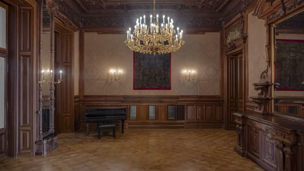 Ornate wooden room with a large chandelier, parquet floors, wall sconces, a grand piano, and a tapestry hanging on the wall.