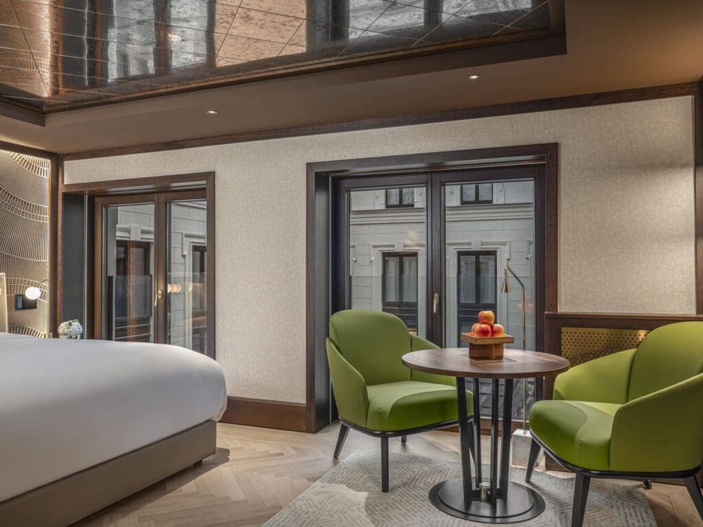 An Almanac Room features a large bed, two green armchairs around a small round table with fruit, and floor-to-ceiling windows overlooking another building.