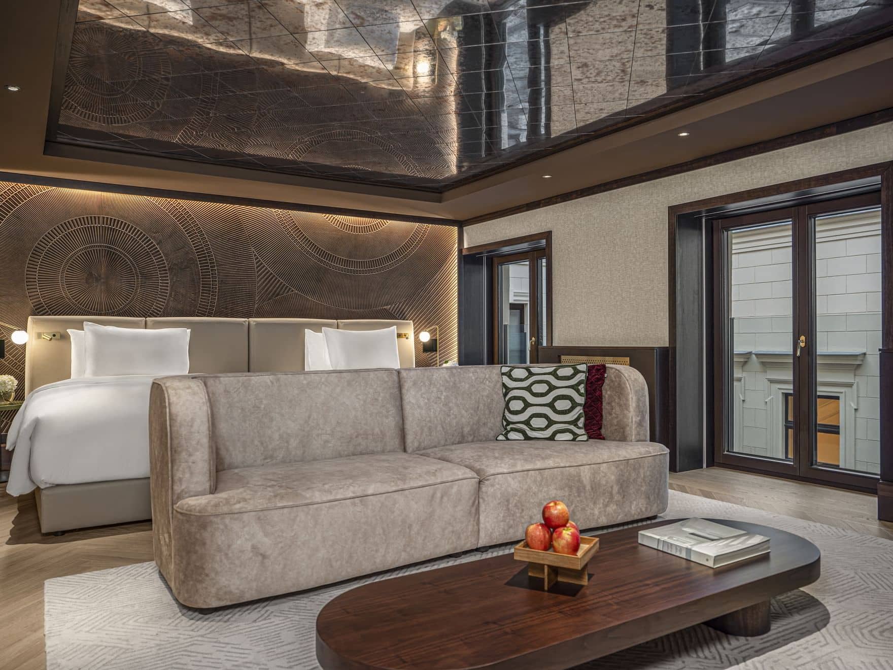 Modern Junior Suite featuring a large bed, beige sofa, low wooden coffee table with apples and a book, and ceiling mirrors reflecting the spacious interior.