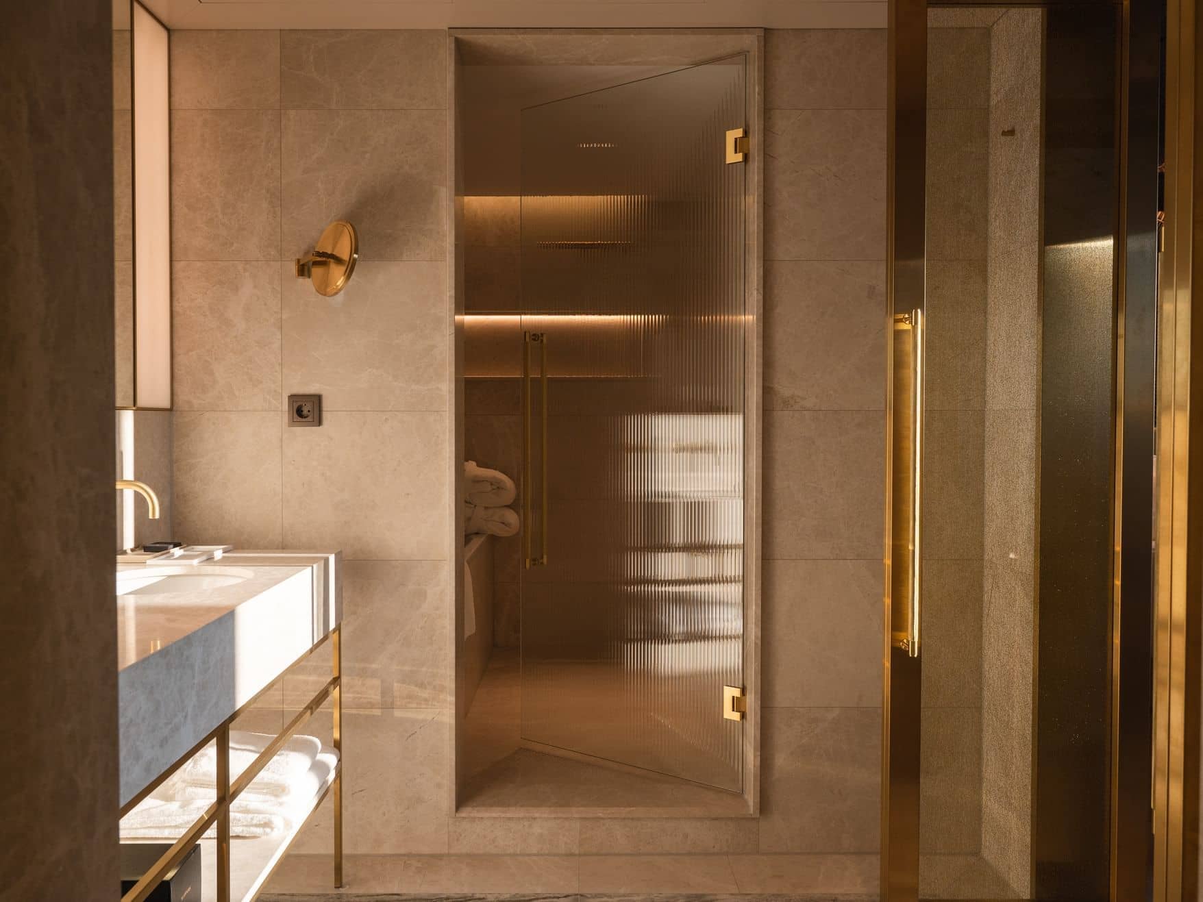 A modern Junior Suite bathroom features a glass shower door, gold fixtures, a marble sink countertop, and beige tiled walls, all illuminated by warm lighting.