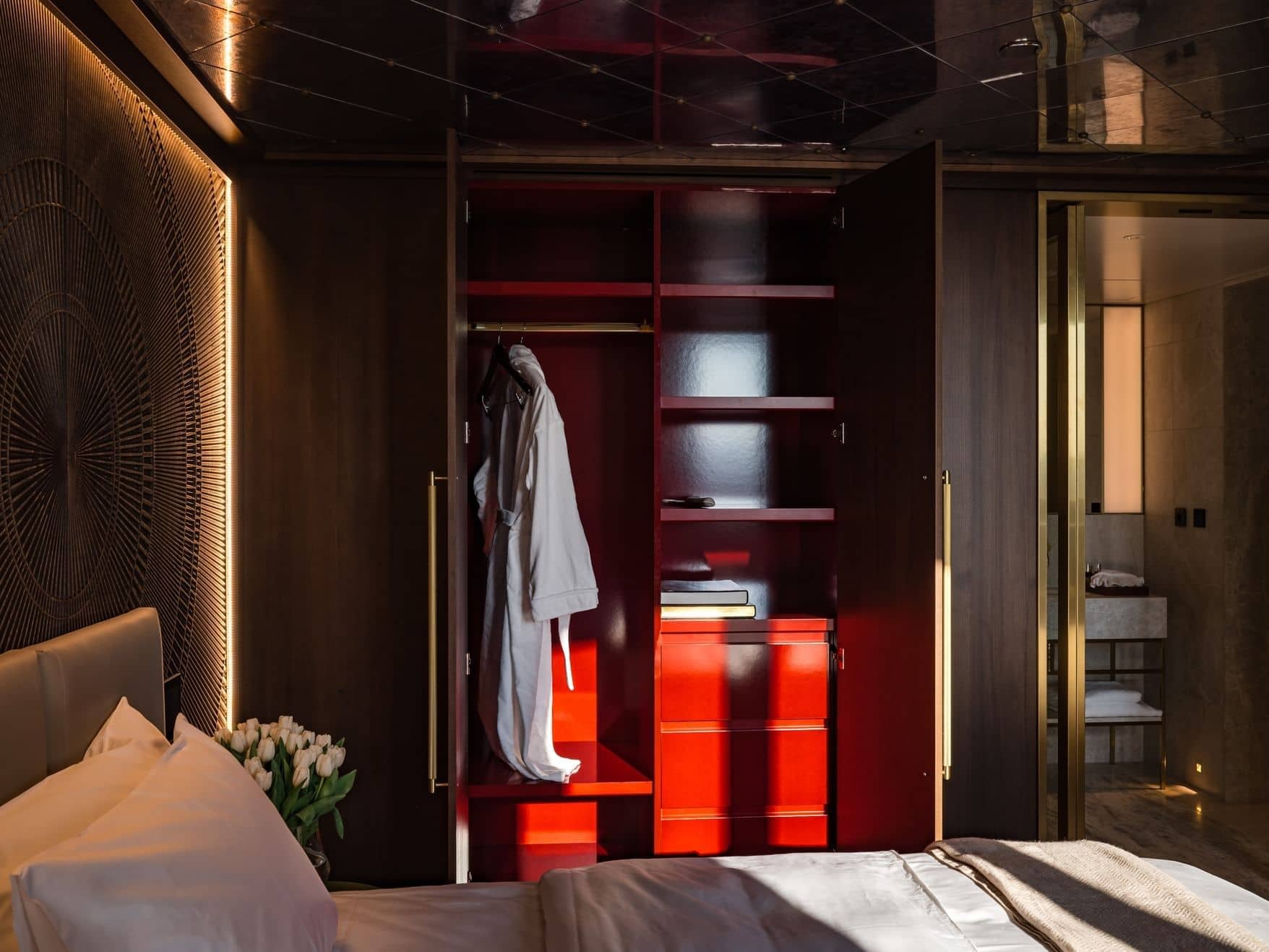 Open wardrobe in the Junior Suite with a robe hanging inside, red shelves, and a few folded items, situated next to a neatly made bed and an adjacent bathroom.
