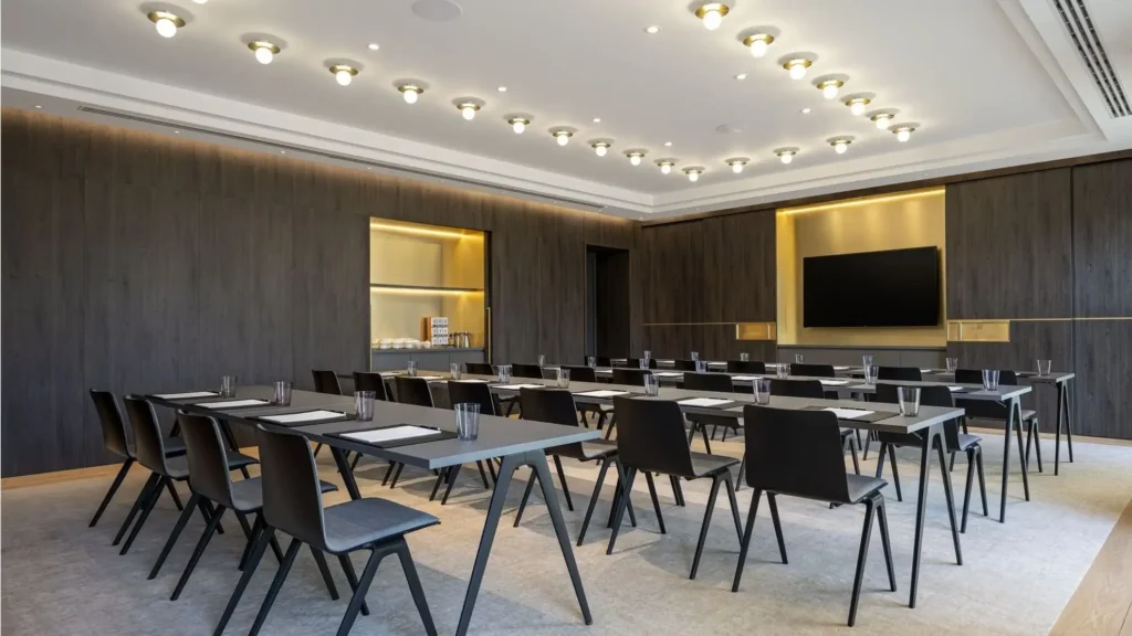 A modern conference room with rows of black chairs and tables facing a wall-mounted screen, under ceiling lights, with glasses and notepads on the tables.