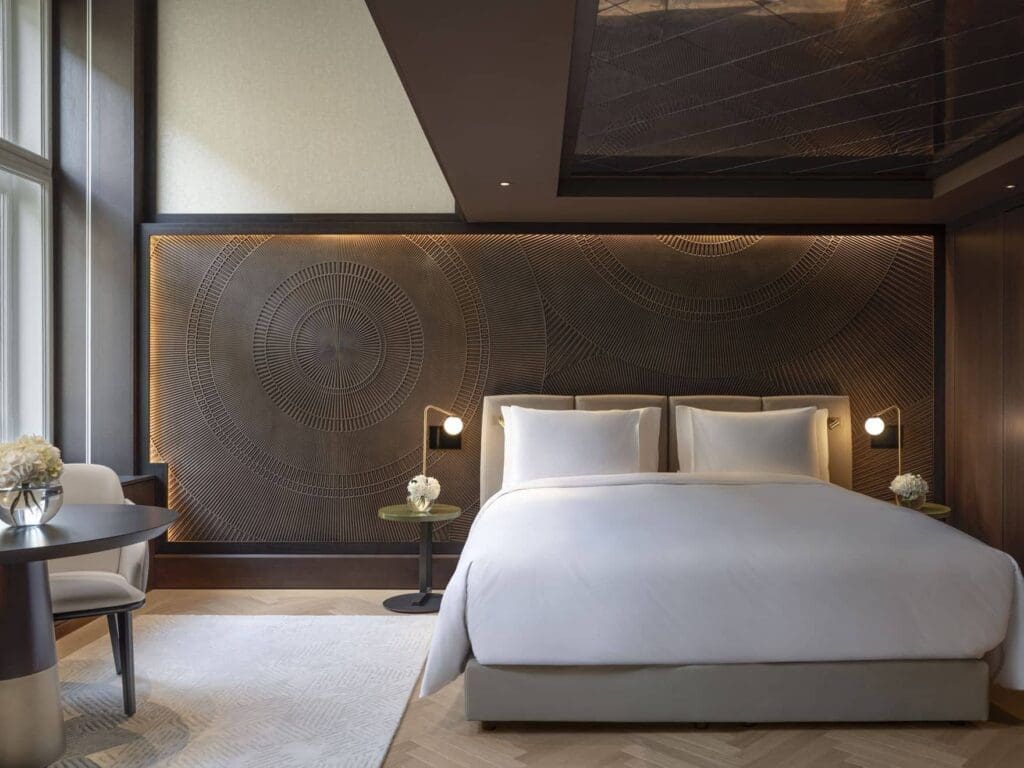 Modernes Hotelzimmer mit einem großen Bett, einer gemusterten Akzentwand, Nachttischen mit Lampen, einem runden Tisch mit Stühlen und bodentiefen Fenstern.