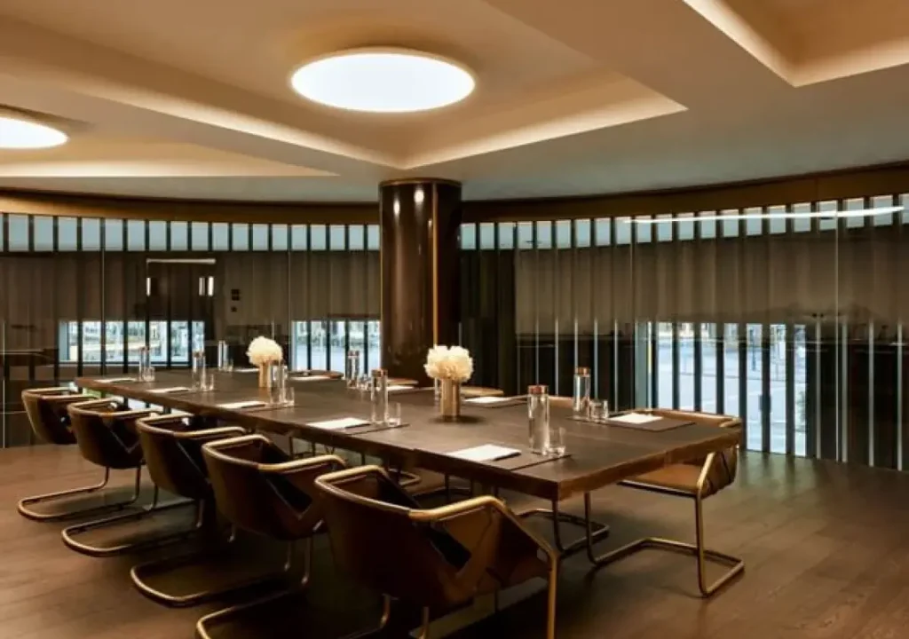 Modern conference room ideal for meetings and events, featuring a long table, brown chairs, notepads, bottled water, and white flower centerpieces. The space boasts wood flooring and floor-to-ceiling windows with blinds.