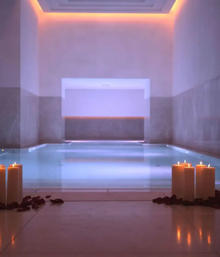 Das Hallenbad im Almanac Palais Vienna erstrahlt in sanftem violettem Licht, umgeben von hohen, brennenden Kerzen und Rosenblättern, die elegant am Beckenrand verstreut sind.