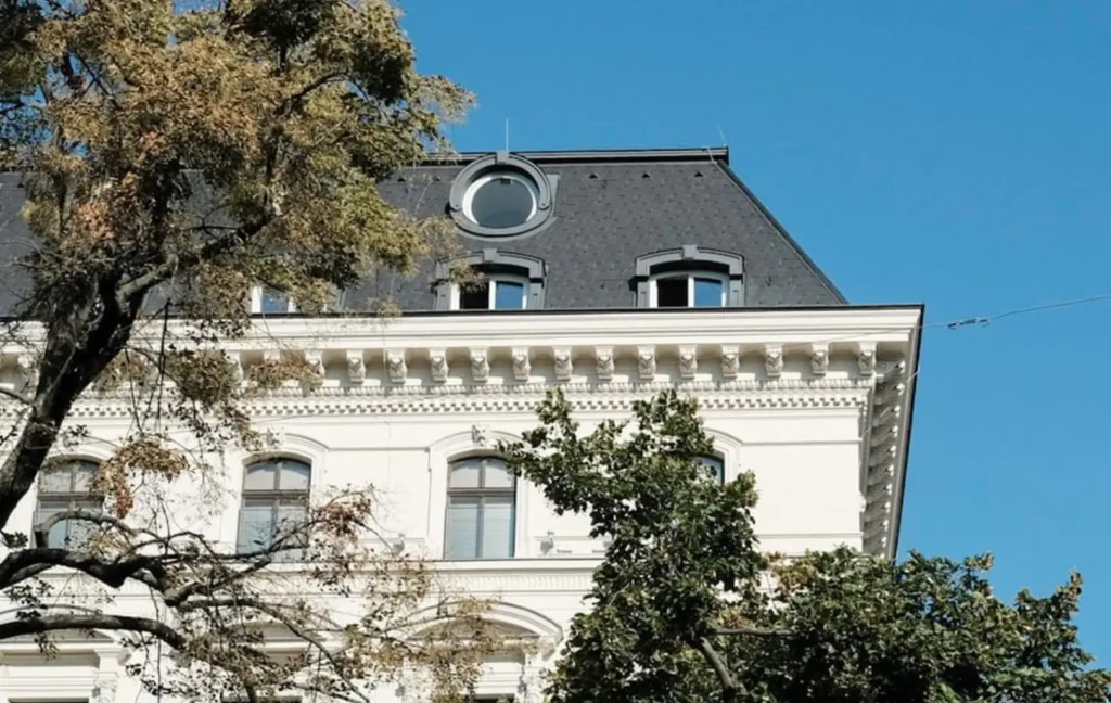 The upper floors of a white historic building with arched windows and a round attic window, partially obscured by leafy trees, under a clear blue sky.