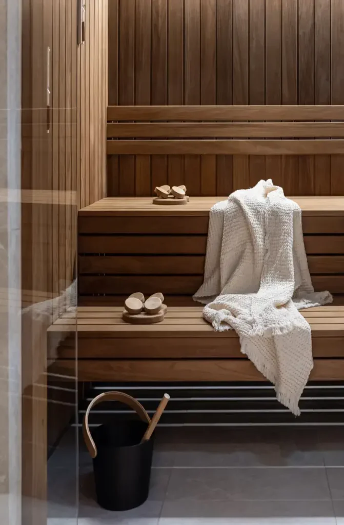 A wooden sauna interior at Almanac Palais in Vienna features a bench with a knit towel draped over it, wooden slippers, and a black bucket with a ladle resting on the floor.