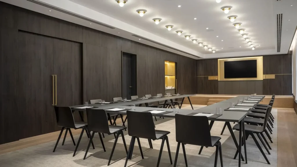 A modern conference room with two rows of black chairs and tables, notepads and glasses on each table, a large screen on the wall, and ceiling lights overhead.
