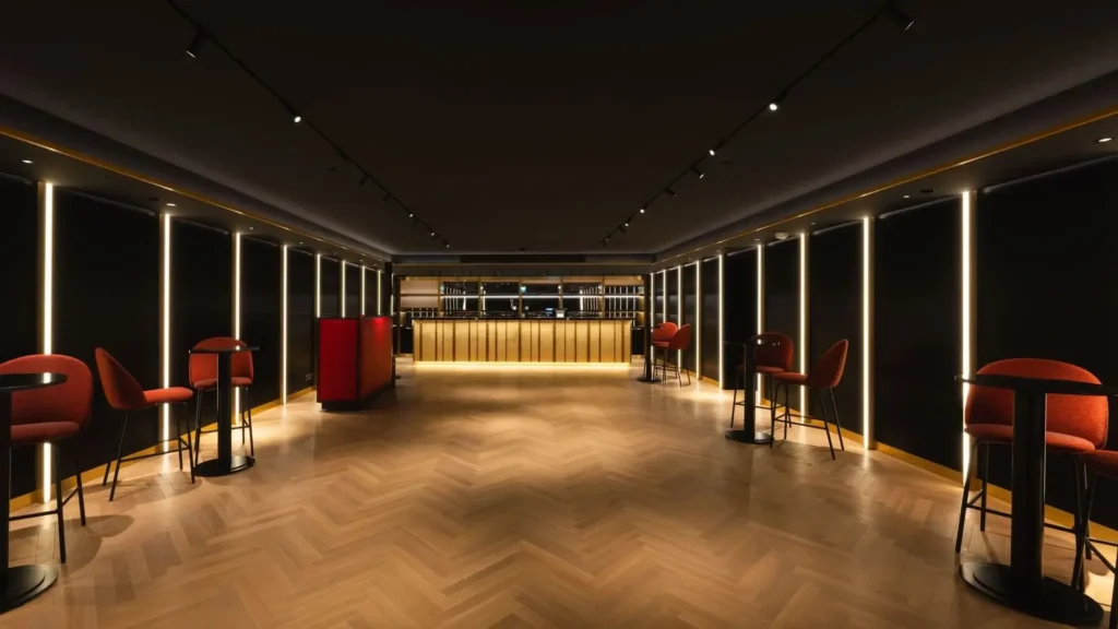 A modern, dimly lit bar with a central counter, high tables with red chairs, herringbone wood flooring, and vertical light strips on dark walls.