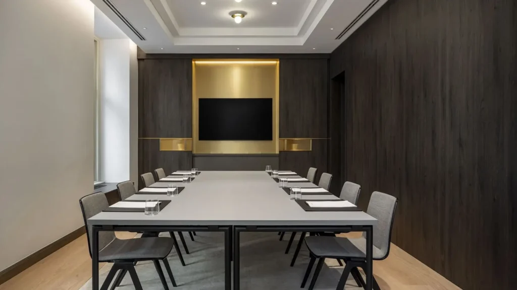 Modern conference room with a rectangular table, ten chairs, notepads, glasses of water, a mounted screen, and dark wood paneled walls.