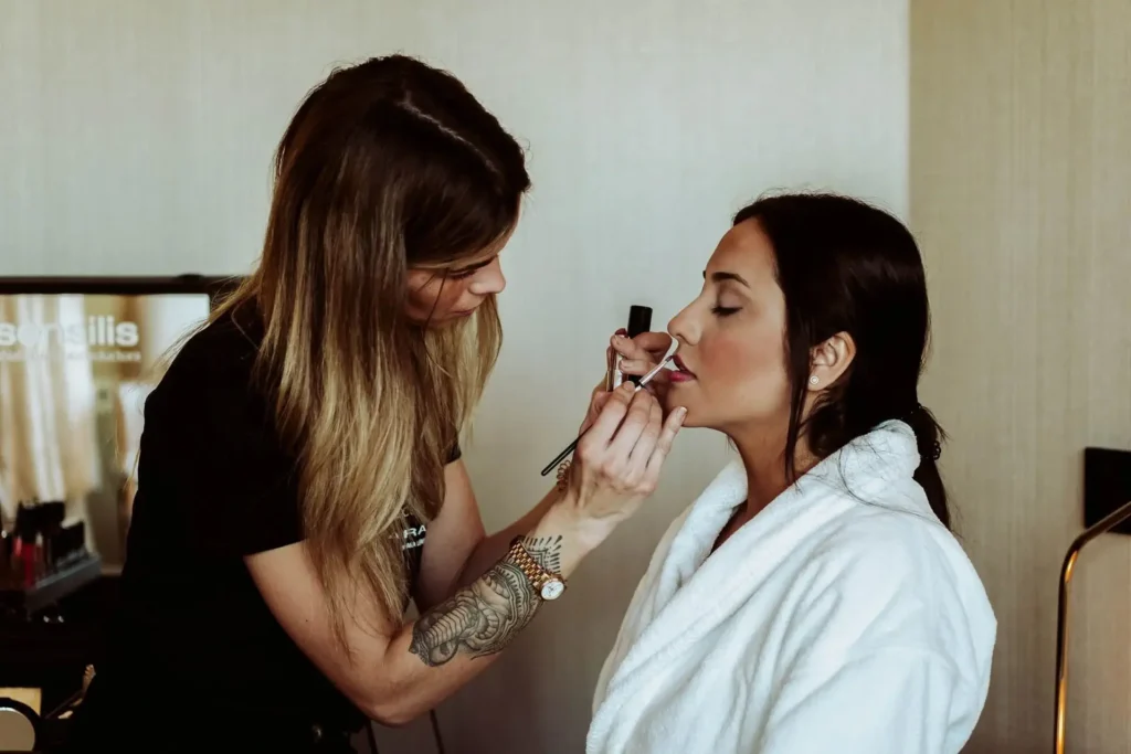 A makeup artist applies lipstick to a woman wearing a white robe, who is seated with her eyes closed.