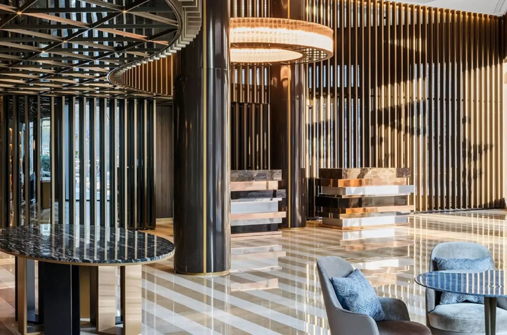 Modern hotel lobby with striped marble floors, dark vertical slat walls, circular chandeliers, and a seating area with blue chairs and a round table.