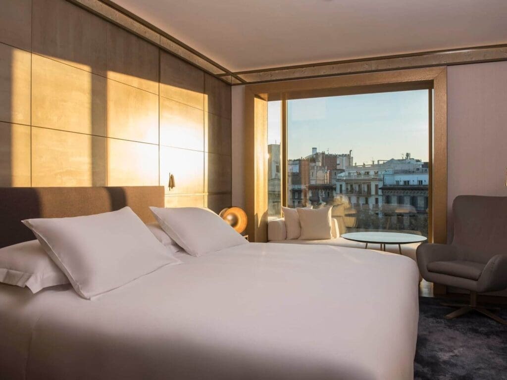 A modern Almanac Suite featuring a large bed, armchair, small table, and floor-to-ceiling window overlooking city buildings at sunset.