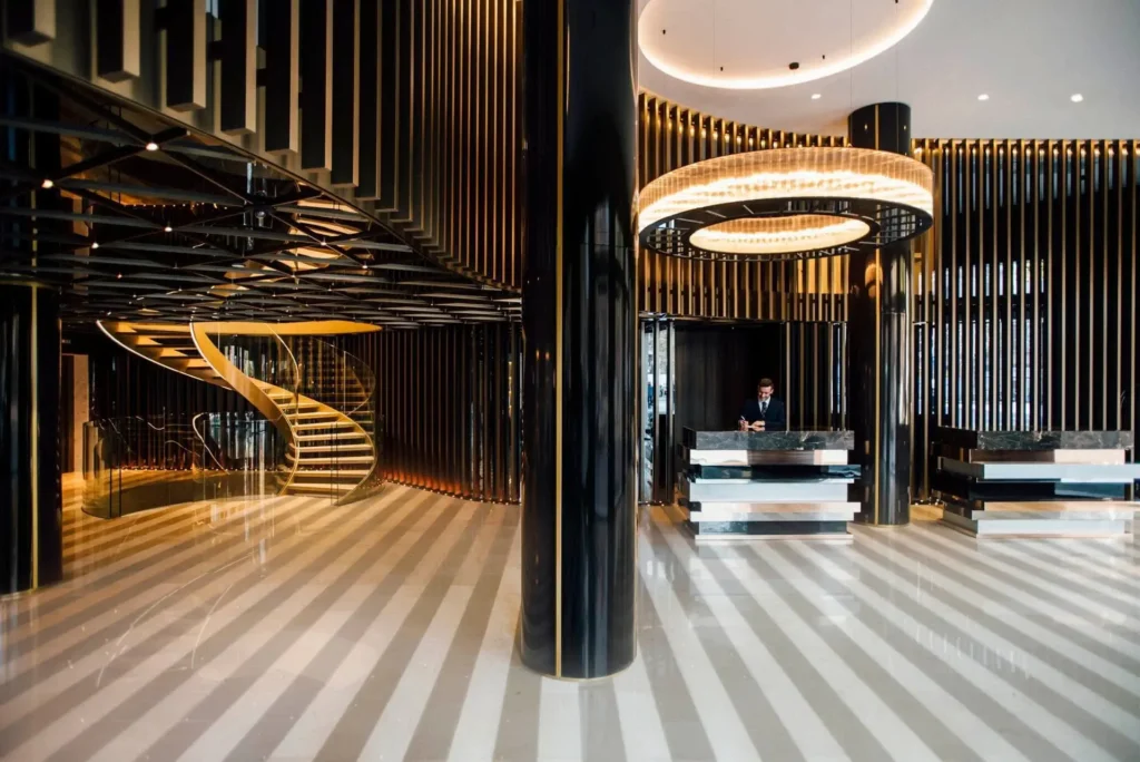 Modern hotel lobby with black columns, striped flooring, a spiral staircase on the left, and a reception desk with a person standing behind it on the right.