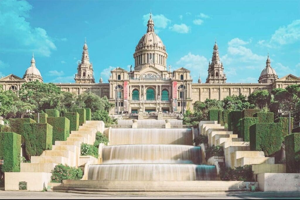 The Museu Nacional d'Art de Catalunya (MNAC) is a large historic building with a central dome and two towers, fronted by terraced gardens, staircases, and cascading fountains under a clear blue sky.