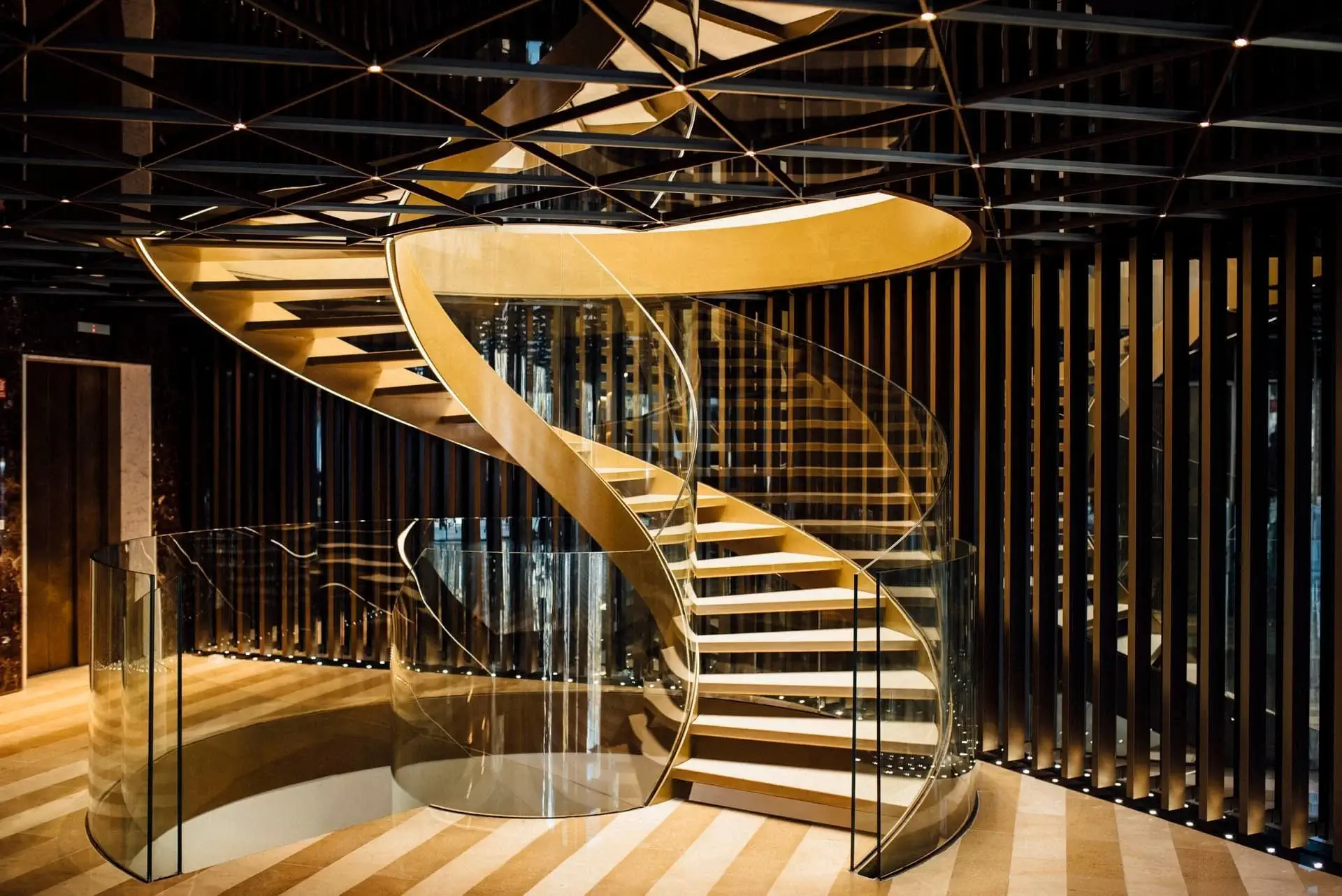 Una escala de cargol moderna amb esglaons i barana de color daurat, barreres de vidre i un terra de fusta a ratlles en un espai interior contemporani.