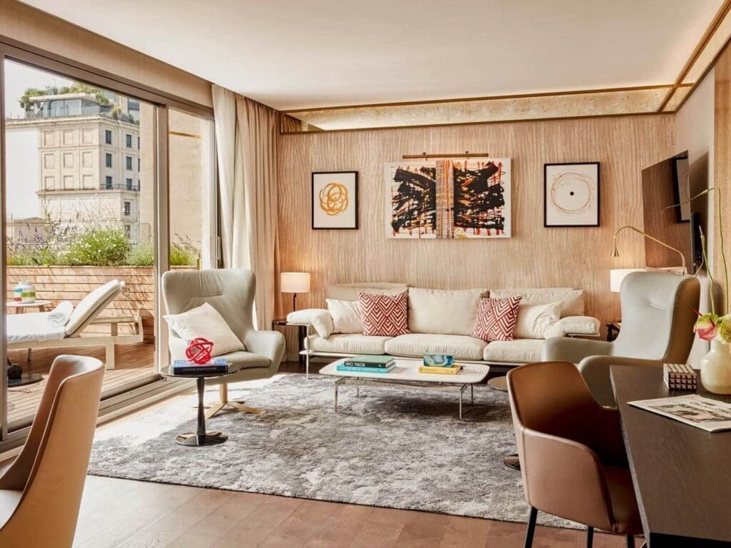 A modern Grand Penthouse Suite living room with a sofa, armchairs, artwork, and a rug, featuring floor-to-ceiling windows that open to a balcony with outdoor seating and breathtaking city views.