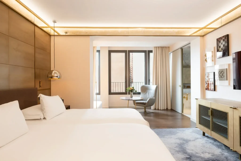 Modern accessible room with two double beds, a chair by the window, a small table with flowers, wall art, and an open doorway to an accessible bathroom.