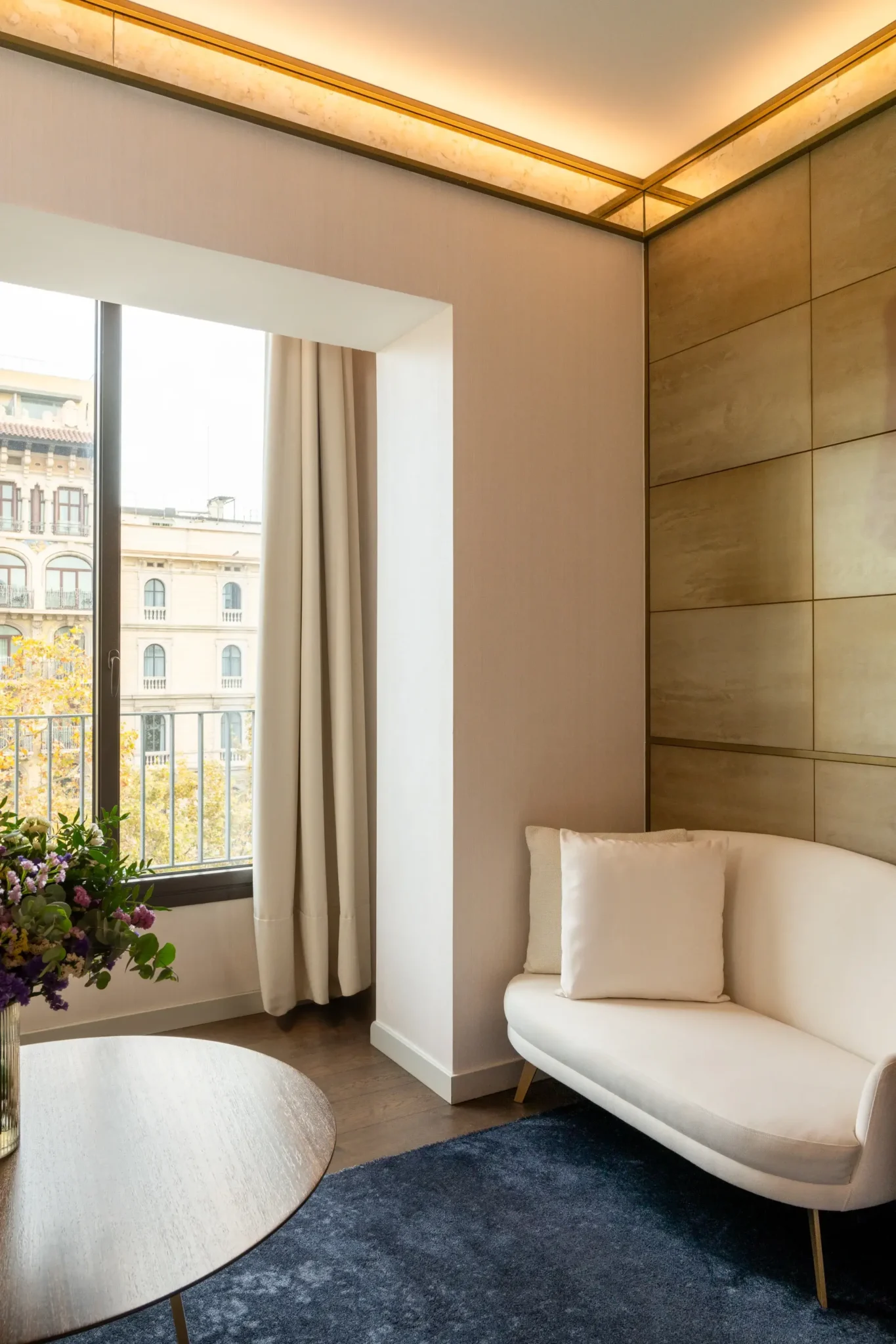 Una pequeña y moderna Sala del Almanaque con un loveseat blanco, una alfombra azul, una mesa redonda de madera con flores, una gran ventana con cortinas color crema que da a un Balcón, y paredes e iluminación en el techo con acentos dorados.