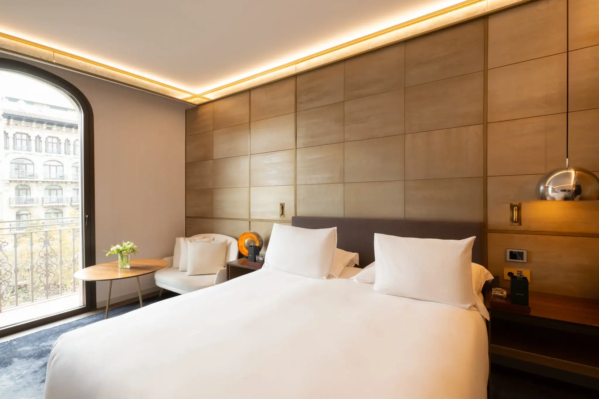 Moderna Habitación Almanaque con una cama grande, dos sillas blancas, una mesa redonda con flores y una amplia ventana o Balcón privado con vistas al paisaje urbano.