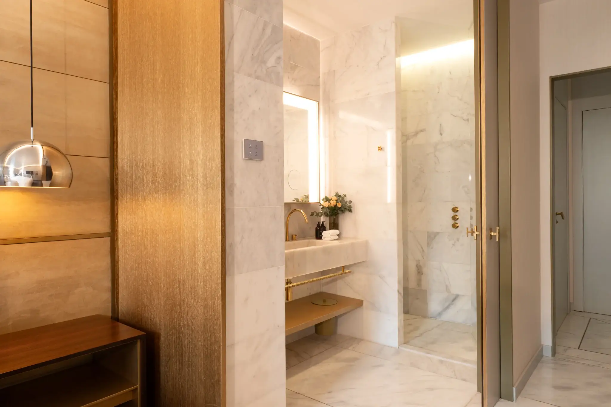 Un moderno cuarto de baño al estilo de la Habitación Almanaque, con paredes y suelos de mármol, una cabina de ducha de cristal, un tocador con espejo y accesorios y detalles en tonos dorados que recuerdan a un elegante refugio de Balcón.