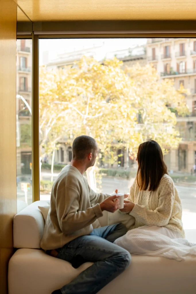 Two people sit on a couch in the Almanac Room by a large window, holding mugs and gazing out at a cityscape of trees and buildings on a sunny day.