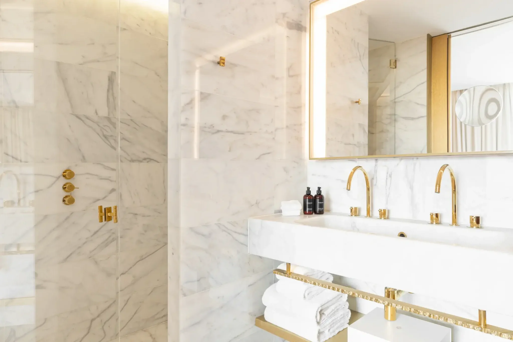 Moderno cuarto de baño con paredes de mármol blanco, grifería dorada, lavabo doble, espejo grande, toallas en estantes y una cabina de ducha de cristal, ideal para una habitación familiar comunicada.