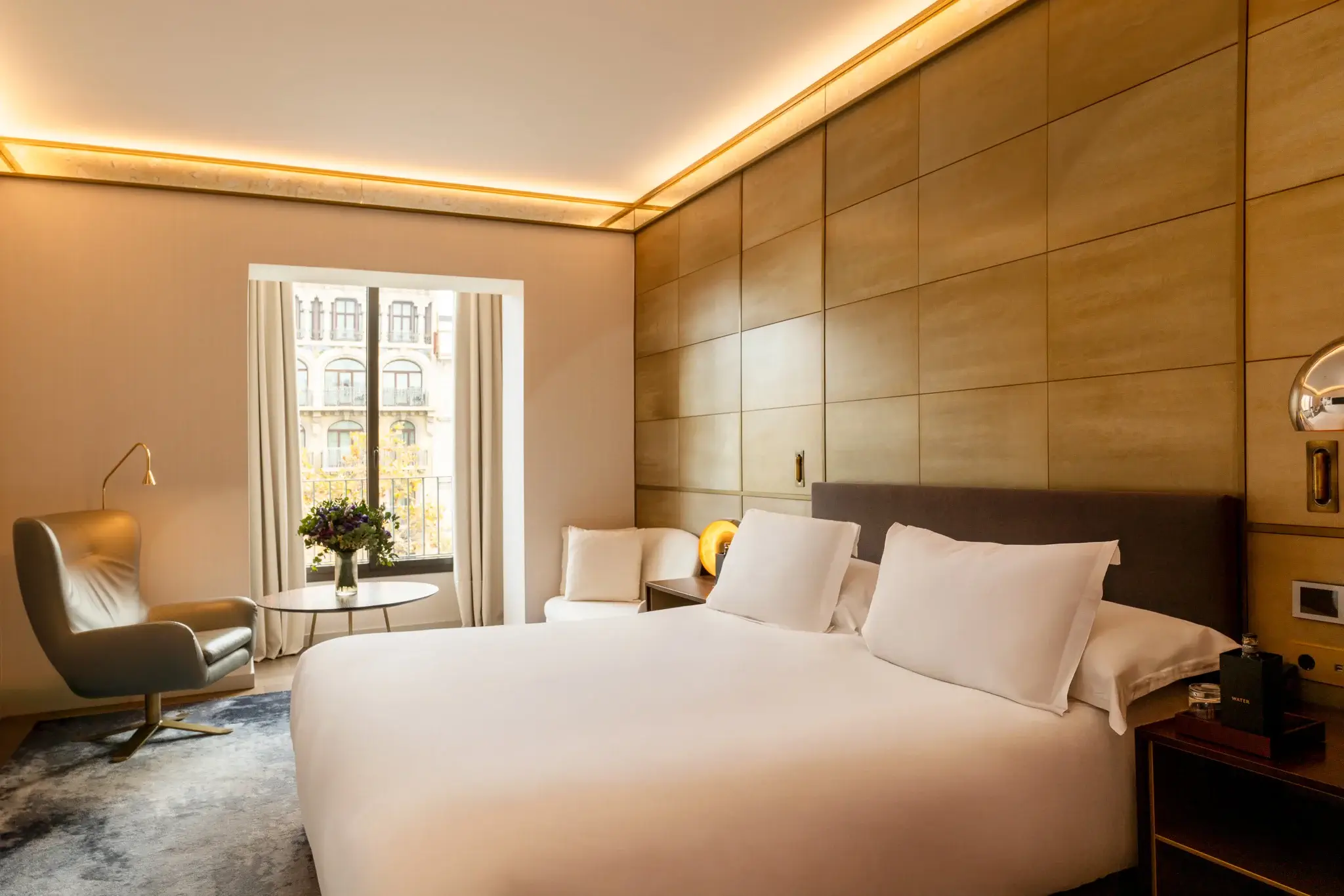 Moderna habitación familiar comunicada con una cama grande, ropa de cama blanca, sillón, mesa auxiliar con flores y una ventana que deja ver los edificios del exterior. La cálida iluminación y las paredes con paneles de madera crean un ambiente acogedor para el disfrute de todos.