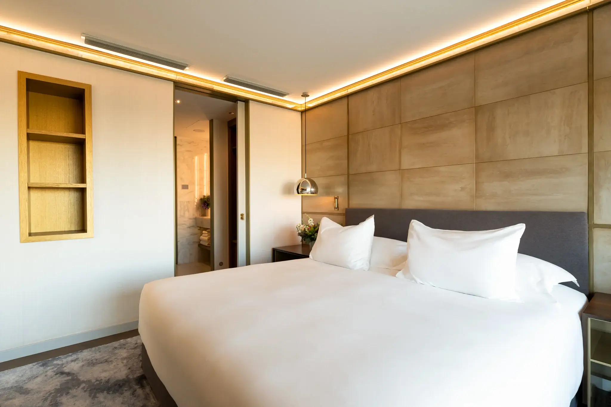 Una moderna habitació d'hotel Almanac amb un llit king size, roba de llit blanca, capçal gris, prestatges integrats, llum penjant i una porta parcialment oberta que condueix a un bany, perfecta per a aquells que busquen la comoditat d'una suite de dues habitacions.