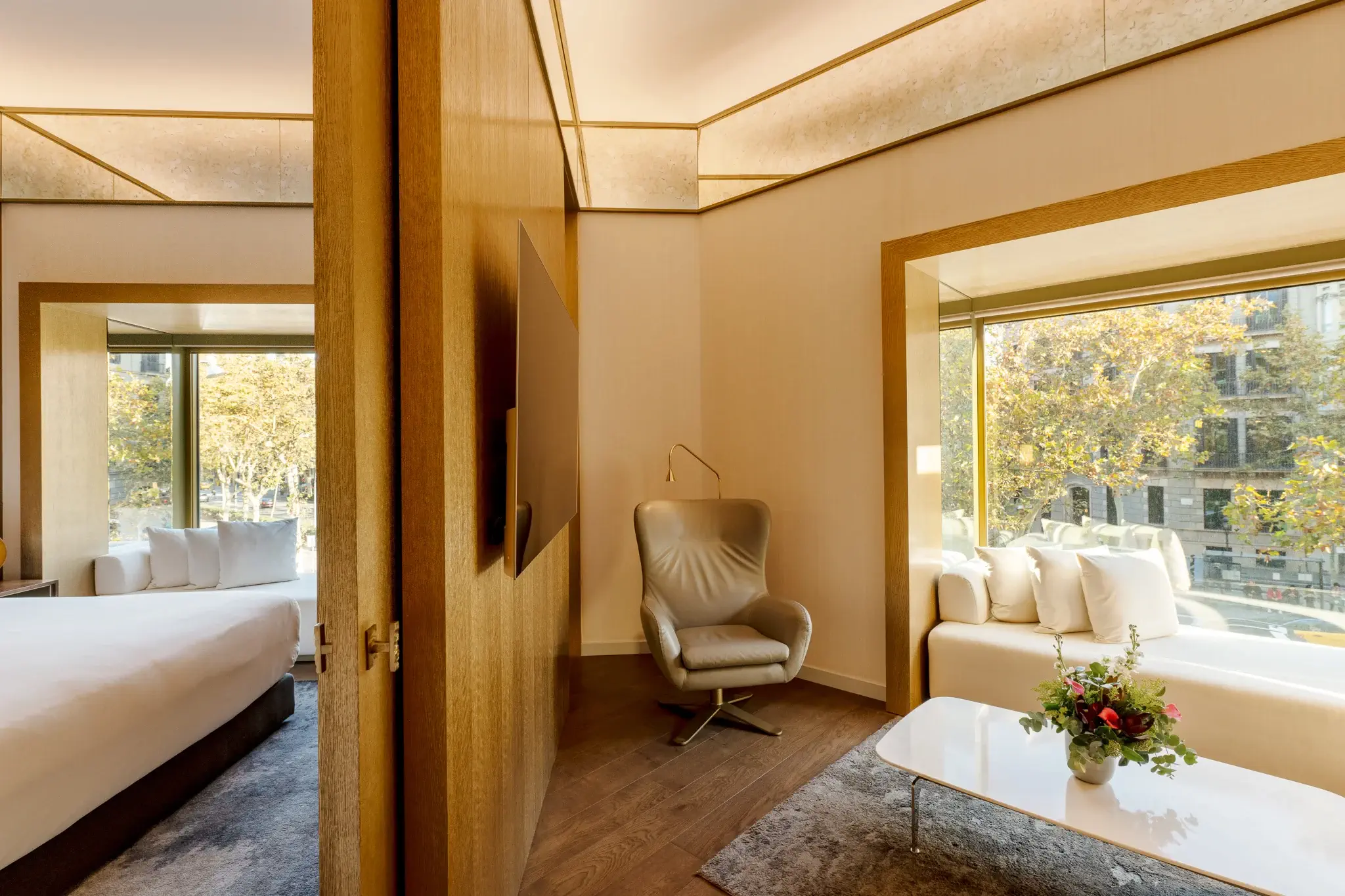 Experimenta la Suite Almanac: una suite d'hotel moderna amb dues habitacions, un llit acollidor, una zona de saló amb butaca i sofà blanc, una taula de centre adornada amb flors, una gran finestra i elegants detalls de fusta.