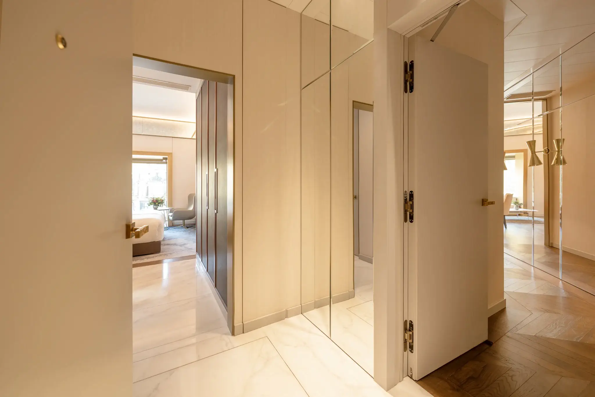 Un passadís amb parets de color crema, terres de marbre, panells de miralls i portes obertes que condueixen a una sala d'estar i a les dues habitacions de la Suite Almanac, totes il·luminades suaument per una llum càlida.