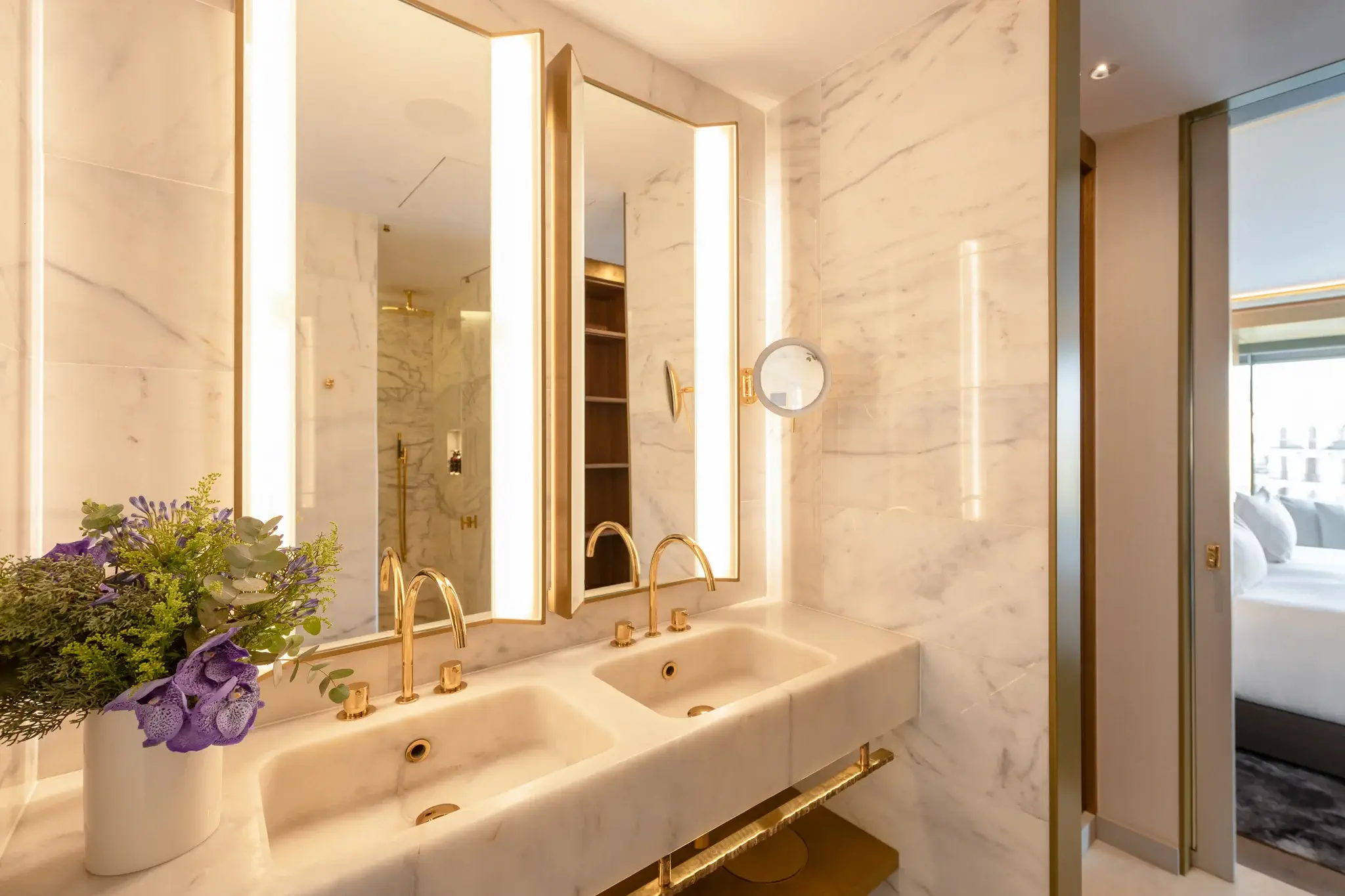 Bany modern amb parets de marbre, lavabo doble amb aixetes daurades, grans miralls il·luminats i un gerro de flors al taulell. La Suite Almanac connecta amb les dues habitacions, amb el dormitori visible a través d'una porta oberta.