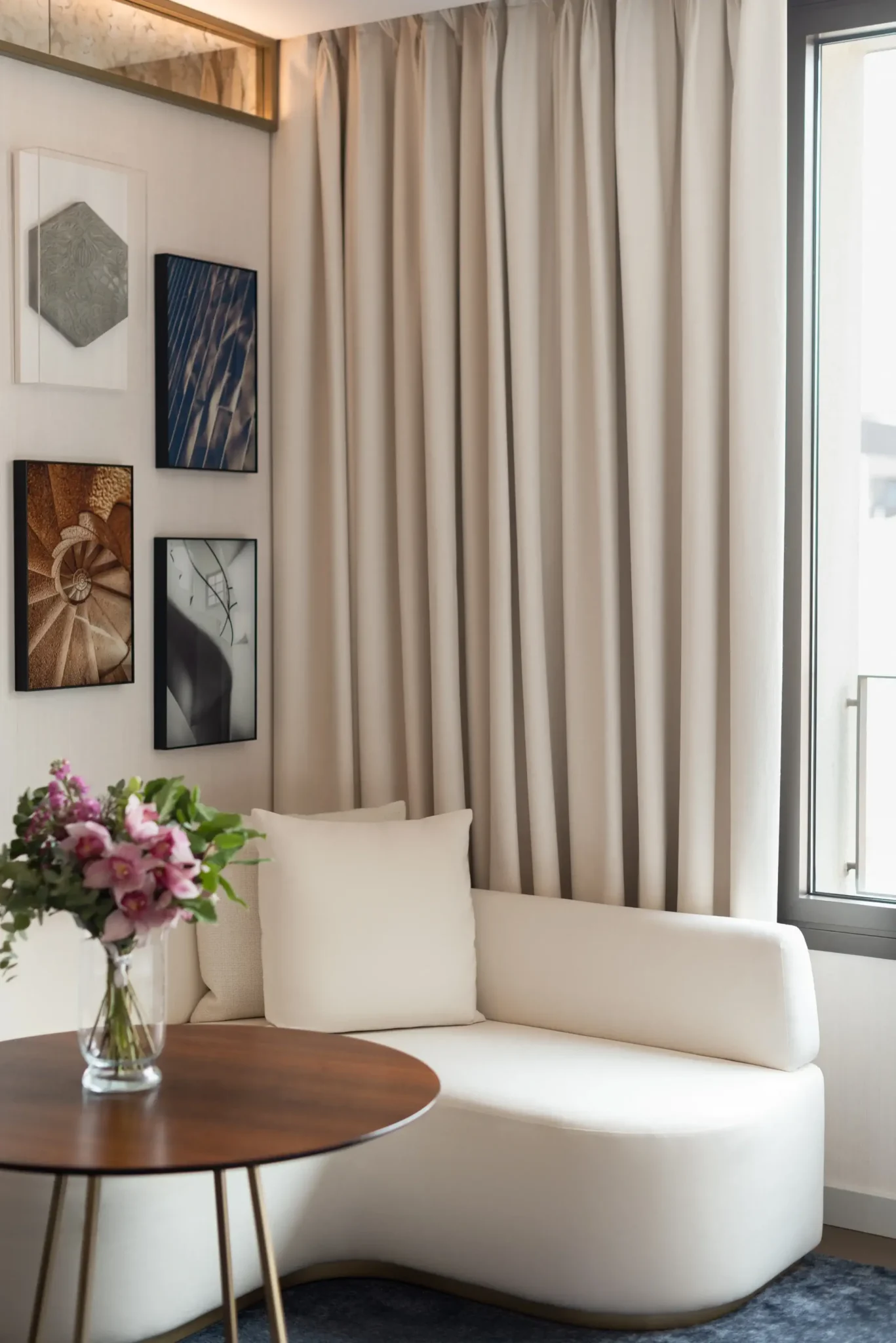 Una sala d'estar moderna amb cantonada de la Habitació Gallery, amb una chaise longue blanca, una taula rodona de fusta amb un gerro de flors, cortines beix i quadres emmarcats.